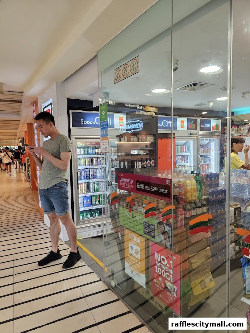A man looks at his phone outside a brightly lit 7-Eleven store with refrigerators full of drinks.