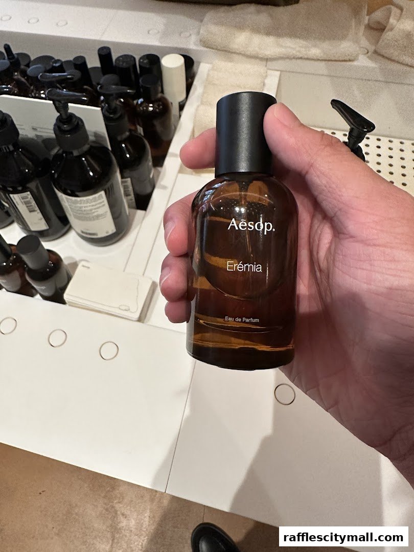 A hand holds an Aesop Eremia Eau de Parfum bottle with other products in the background.