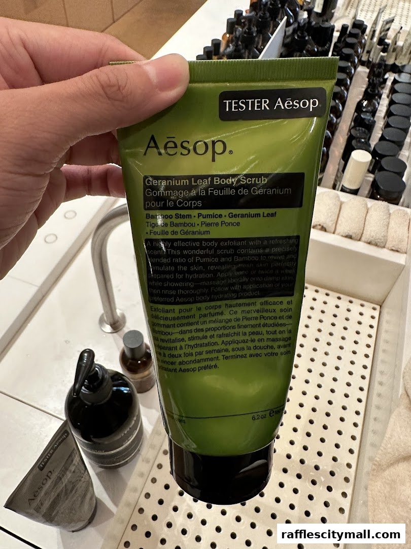 A hand holds an Aesop Geranium Leaf Body Scrub tester tube with other Aesop products in the background.