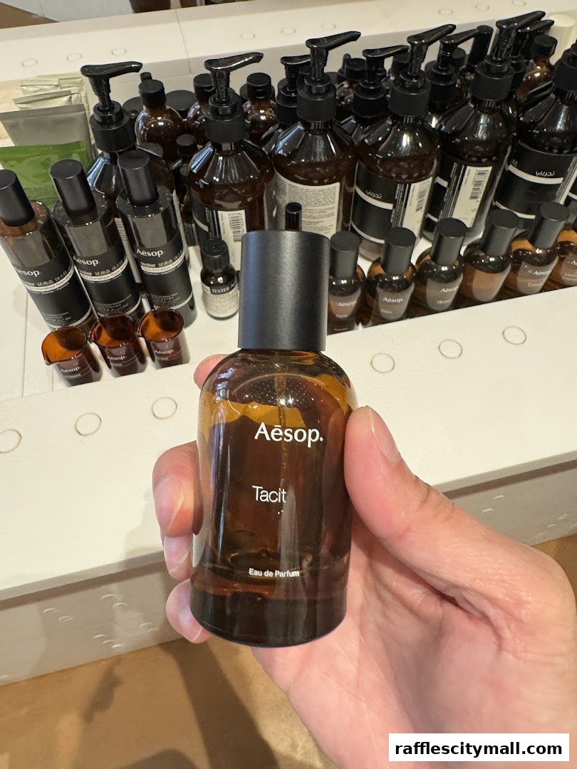 A hand holds an Aesop Tacit Eau de Parfum bottle in front of a display of Aesop products.