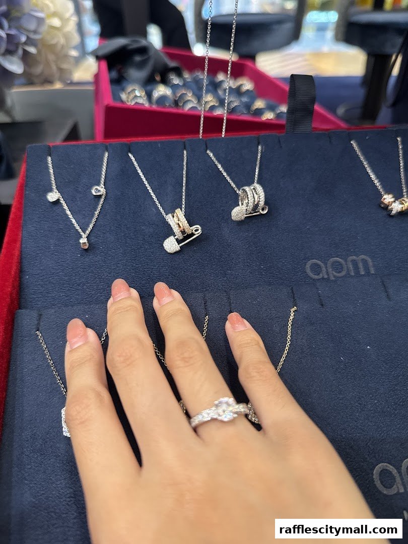 A hand wearing a diamond ring is shown near APM Monaco necklaces displayed on a dark blue velvet stand.