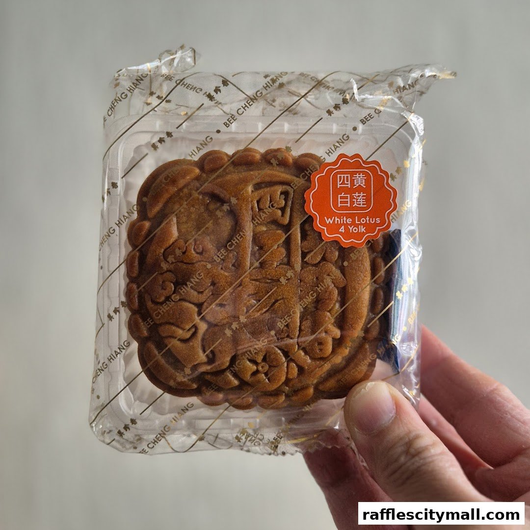 A hand holds a packaged Bee Cheng Hiang White Lotus mooncake with 4 yolks.