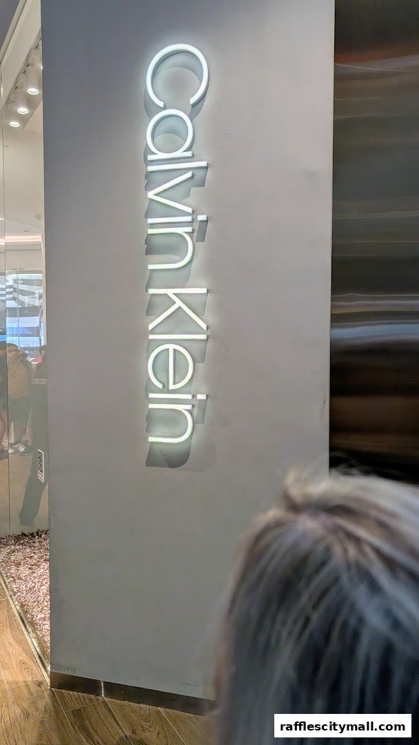 A vertical neon sign displays the Calvin Klein logo on a grey wall.
