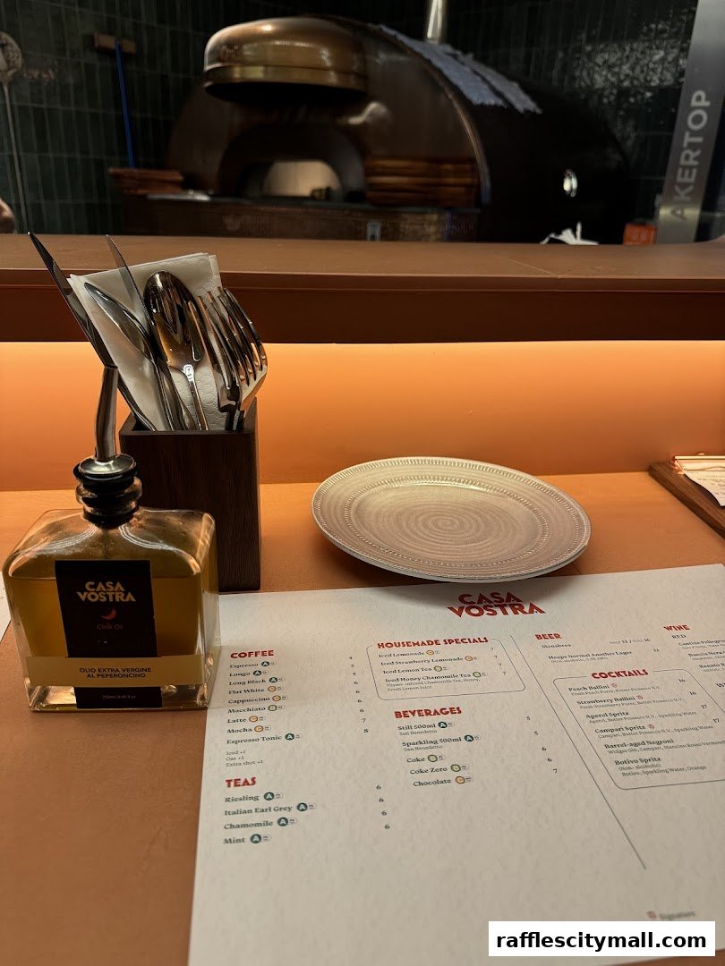 A table setting at Casa Vostra with a menu, olive oil, and cutlery, featuring a pizza oven in the background.