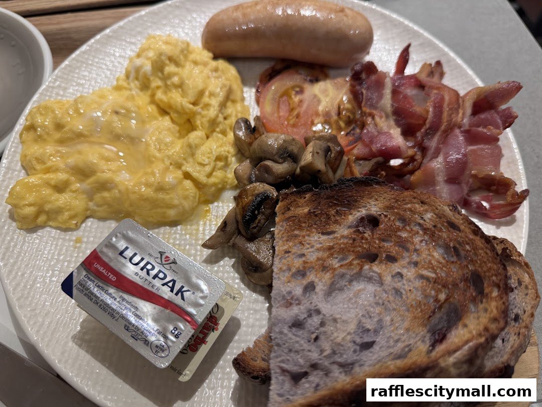 A close-up of a hearty breakfast plate with scrambled eggs, bacon, sausage, mushrooms, tomato, and toast.