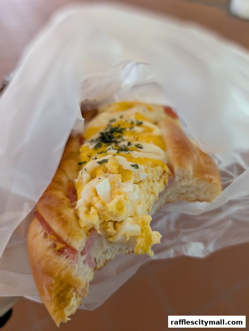 A close-up of a delicious savory pastry filled with egg salad and ham, wrapped in white paper.
