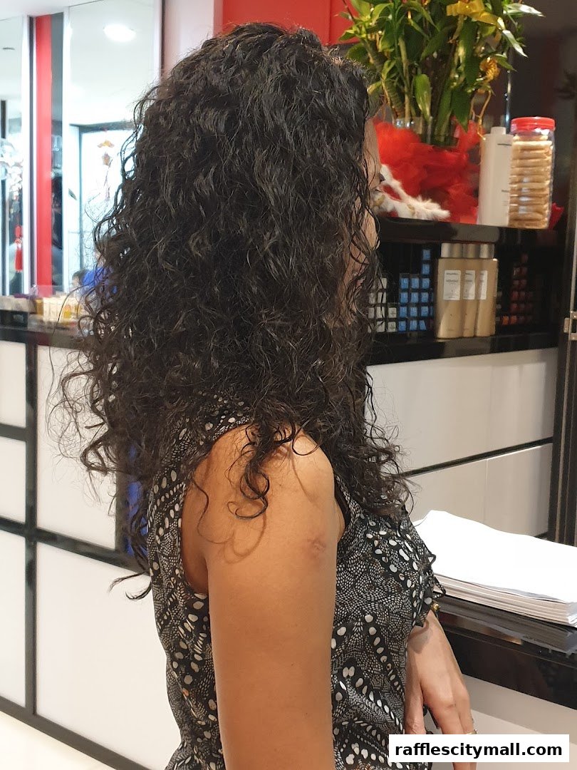 A woman with long, curly dark hair is shown from the side in a salon.