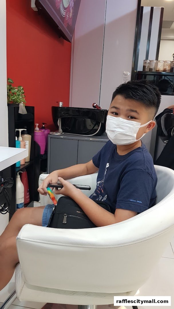 A young boy wearing a mask sits in a salon chair, holding a phone.