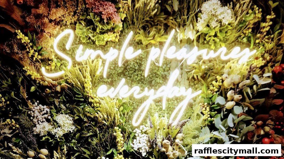 A neon sign reading 'Simple pleasures everyday' is set against a lush backdrop of flowers and greenery.