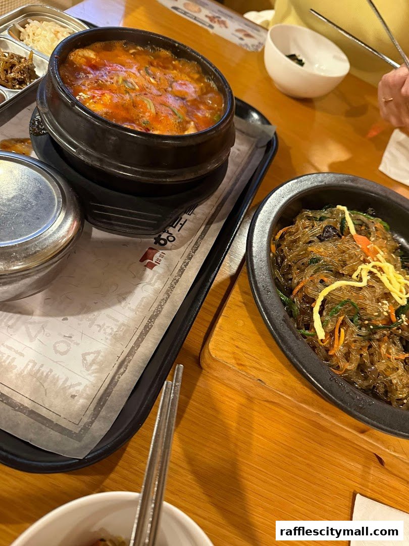 A close-up of Korean dishes, including a bubbling stew and japchae, served on a wooden table.