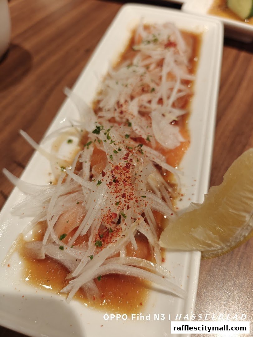 A plate of thinly sliced salmon with onions, chili flakes, and a lemon wedge.