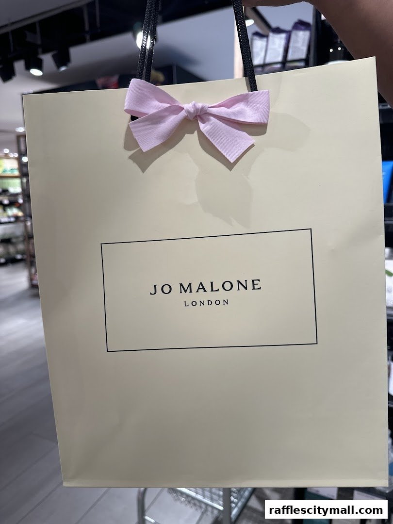 A Jo Malone London shopping bag with a pink bow is held up.