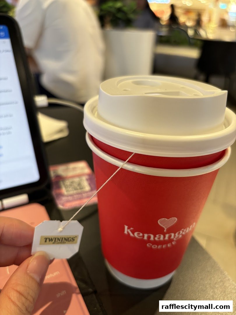 A hand holds a Twinings tea bag next to a red Kenangan Coffee cup.