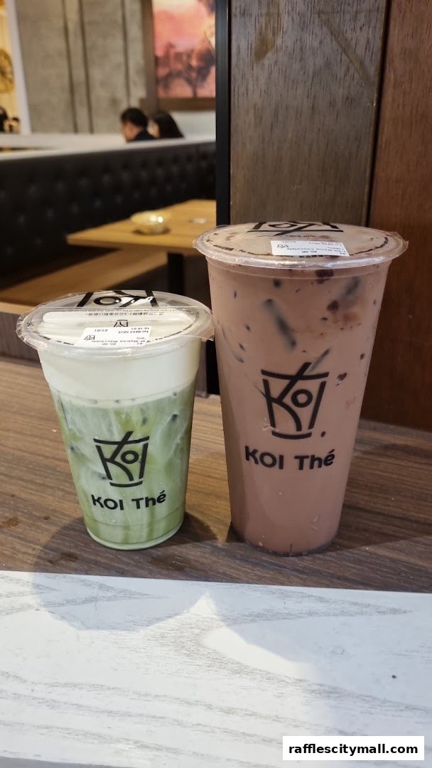 Two KOI Thé drinks, one matcha with cream and one chocolate, are displayed on a wooden surface.