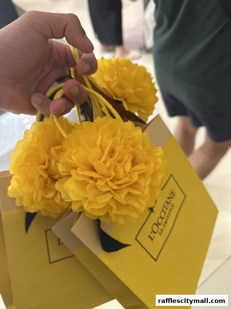 A hand holds yellow shopping bags from L'Occitane decorated with fluffy yellow flowers.
