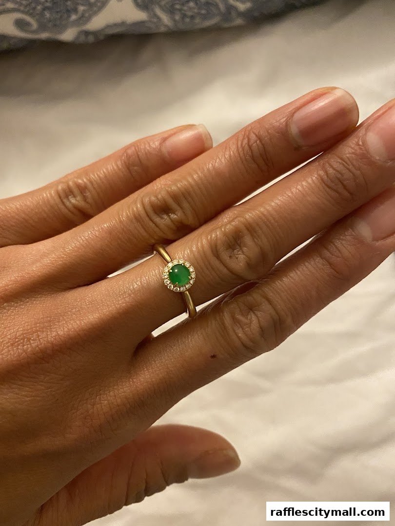A hand wearing a gold ring with a green jade stone surrounded by small diamonds.
