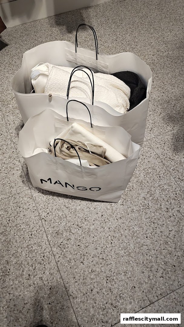 Two white shopping bags from MANGO filled with clothing items on a speckled floor.
