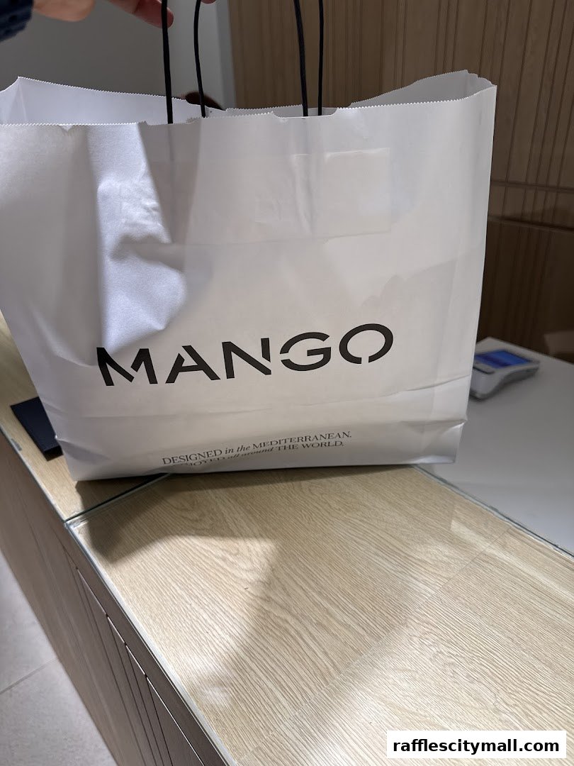 A white shopping bag with the MANGO logo and tagline on a wooden counter.