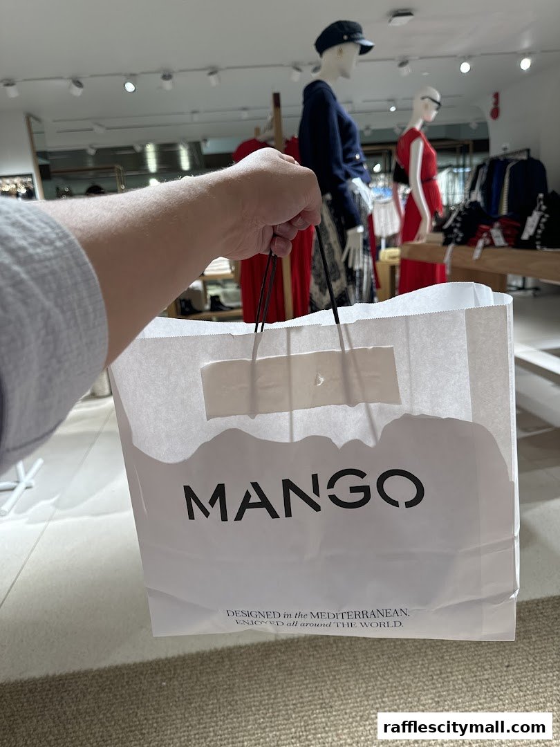 A hand holds a MANGO shopping bag inside a store with mannequins displaying clothes.