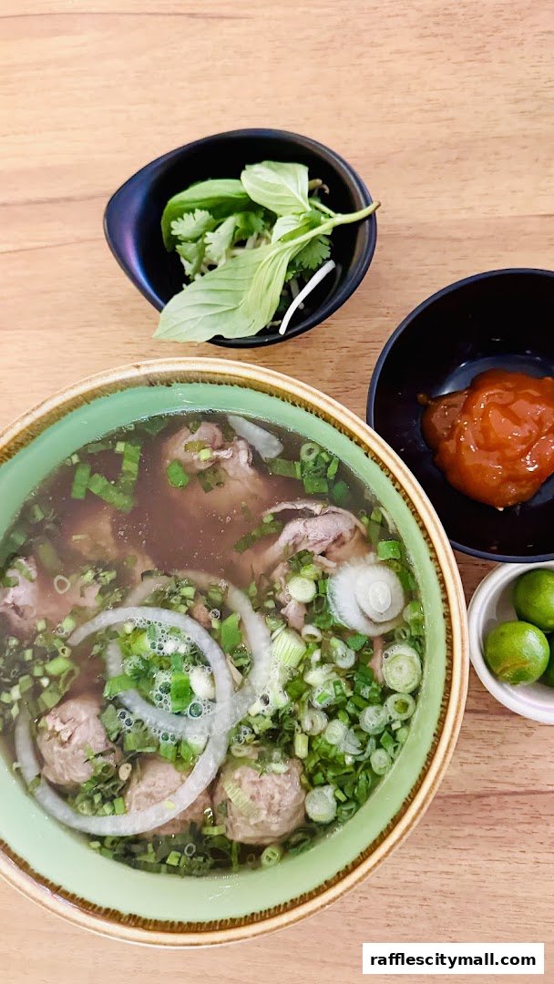 A steaming bowl of pho with beef, meatballs, onions, and herbs, served with lime and chili sauce.