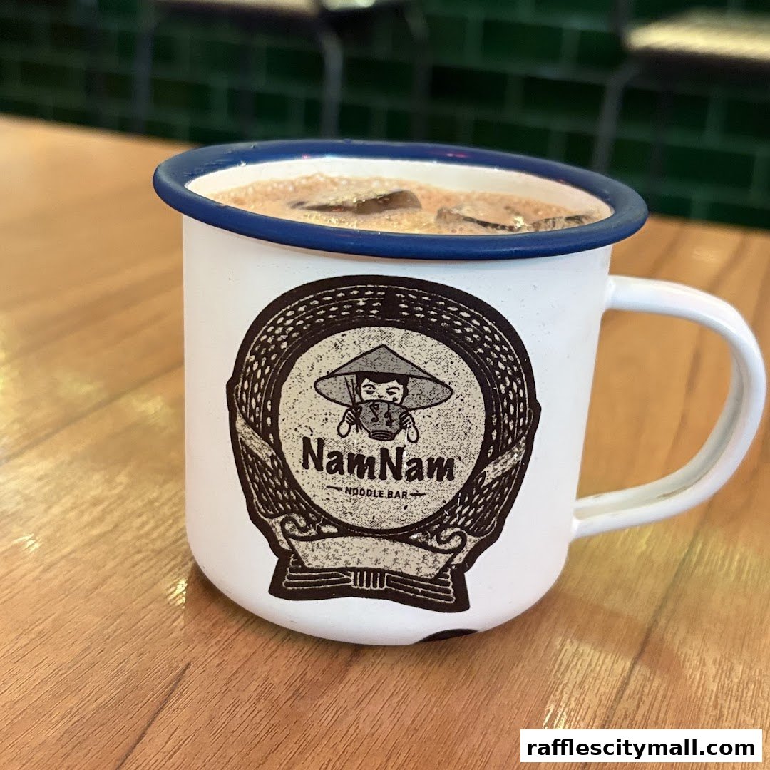 A close-up of a white mug with the NamNam Noodle Bar logo, filled with a beverage and ice.