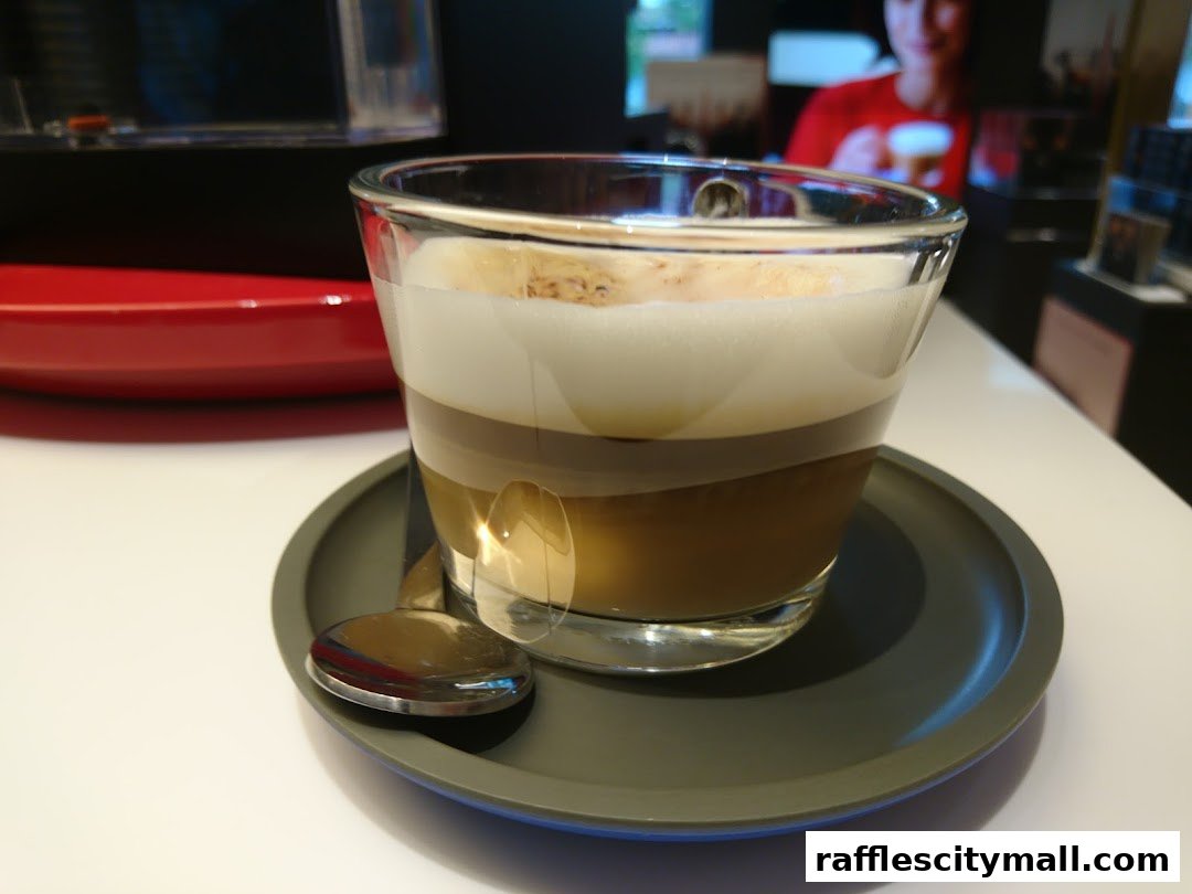 A layered coffee drink with foam served in a glass on a saucer with a spoon.