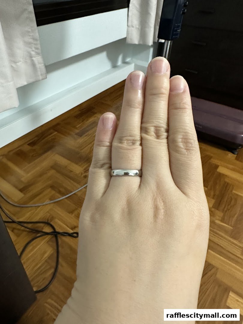 A hand wearing a simple silver band ring, with a wooden floor and curtains in the background.
