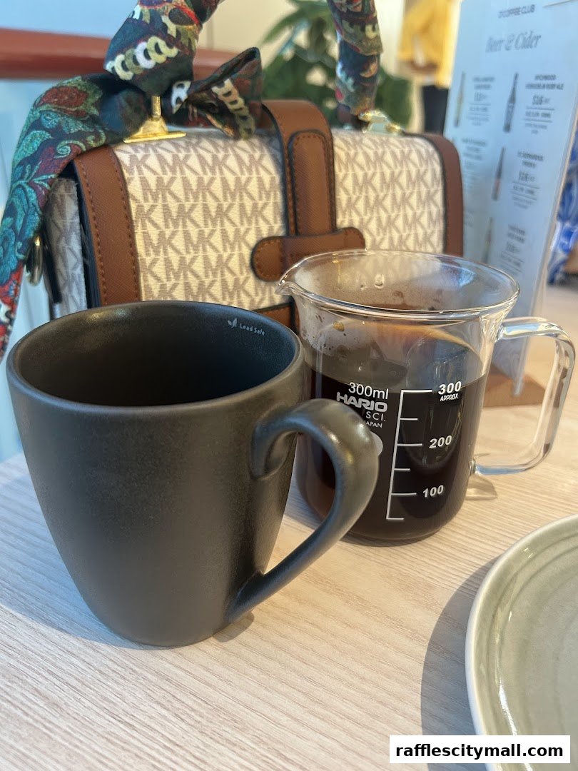 A dark coffee mug and a glass beaker of coffee sit on a wooden table next to a handbag.