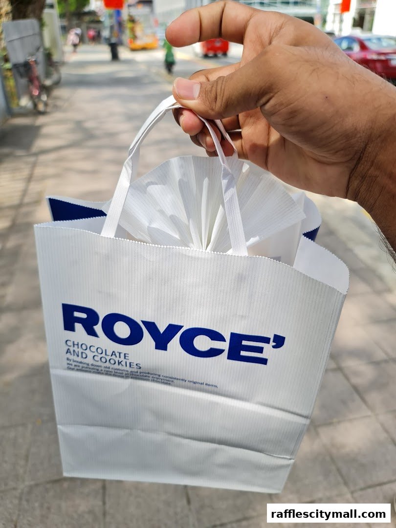 A hand holds a white Royce' shopping bag with blue lettering.