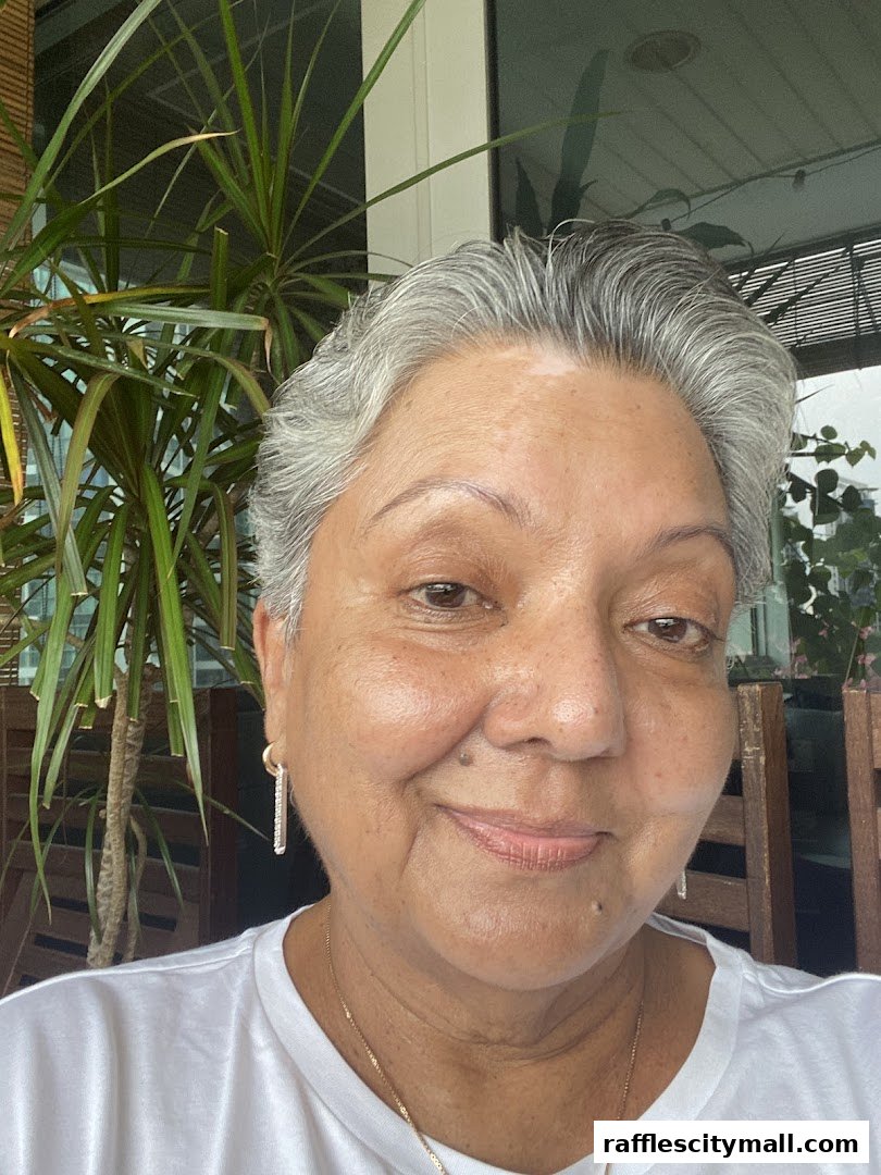 A woman with short grey hair and vitiligo smiles at the camera, with a plant and outdoor seating in the background.