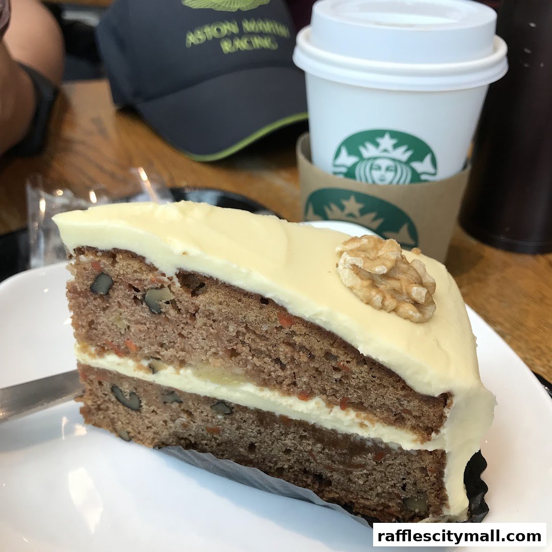 A slice of carrot cake with cream cheese frosting and a walnut on top, next to a Starbucks coffee cup.
