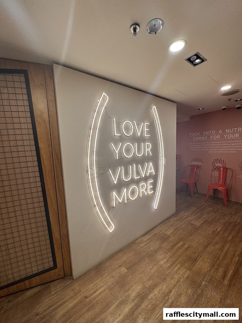A shop interior with a neon sign saying 'LOVE YOUR VULVA MORE' and two red chairs.