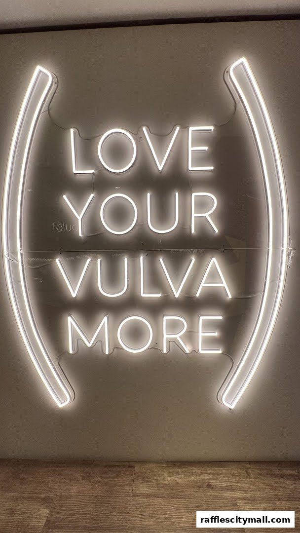 A neon sign with the text 'LOVE YOUR VULVA MORE' is displayed on a wall.