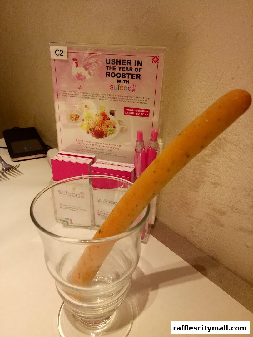 A promotional sign for Sufood's Year of the Rooster Yusheng dish with a breadstick in a glass.