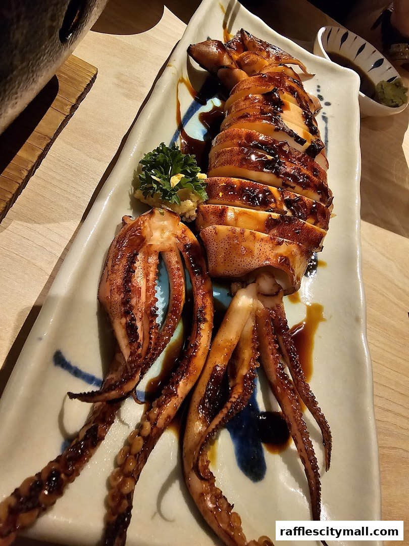 A beautifully presented grilled squid dish with a glistening glaze, served on a ceramic plate with soy sauce and wasabi.