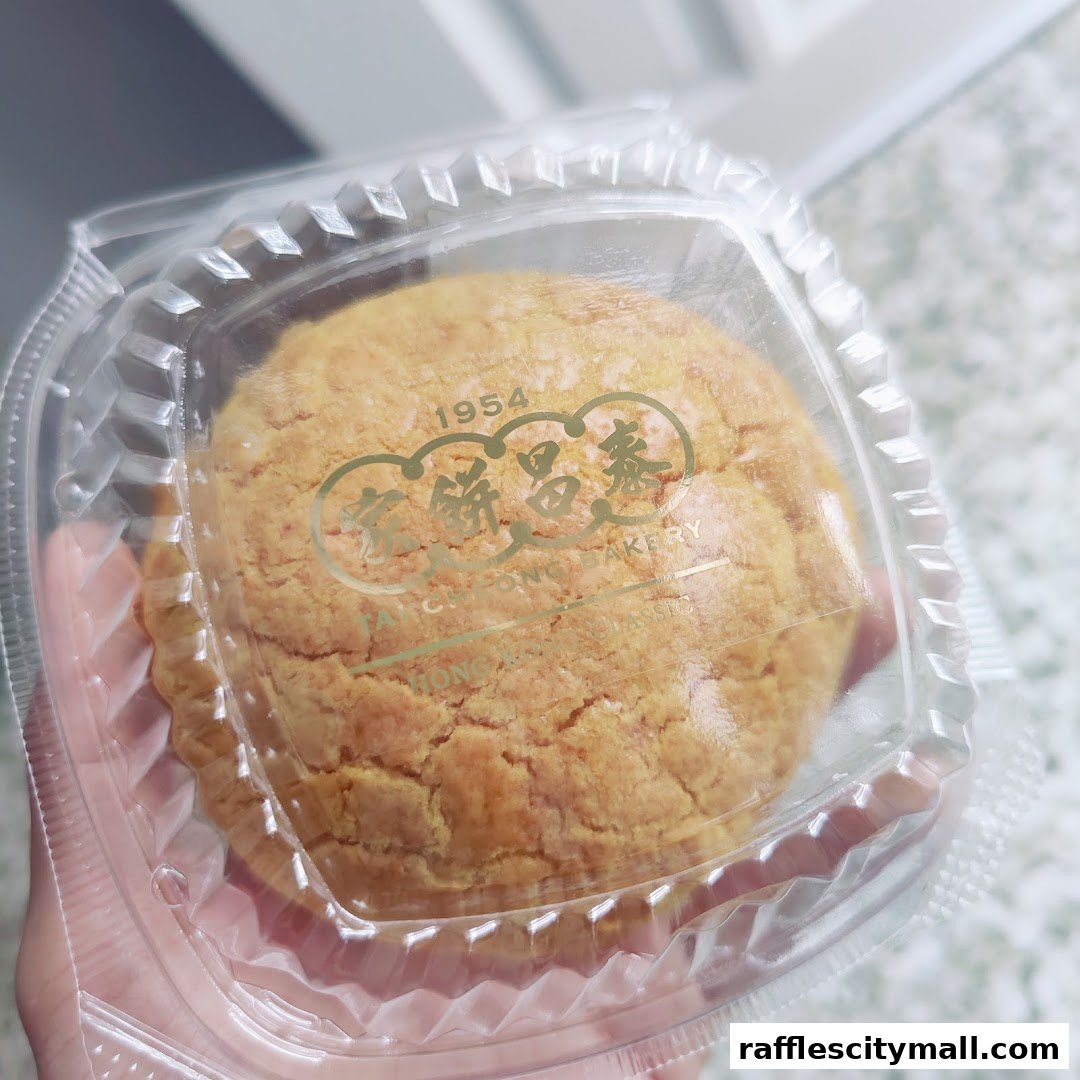 A hand holds a clear plastic container with a golden-brown pastry inside, featuring the Tai Cheong Bakery logo.