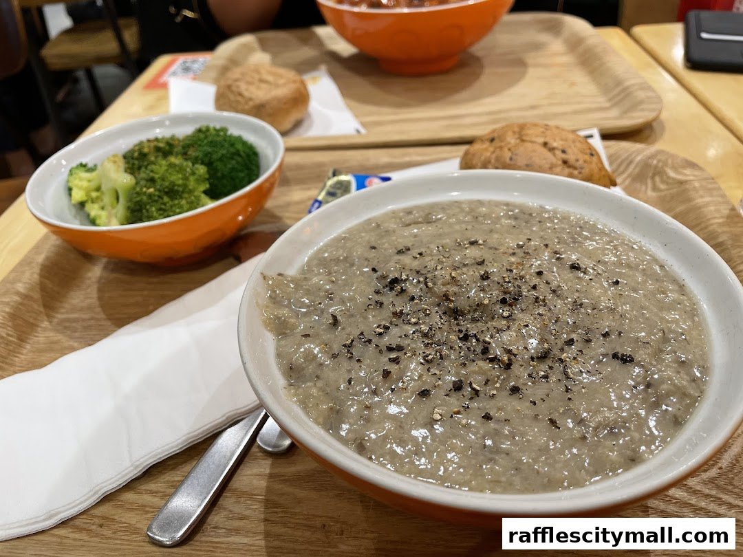 A close-up of a creamy mushroom soup with pepper, served with a side of broccoli and a bun.