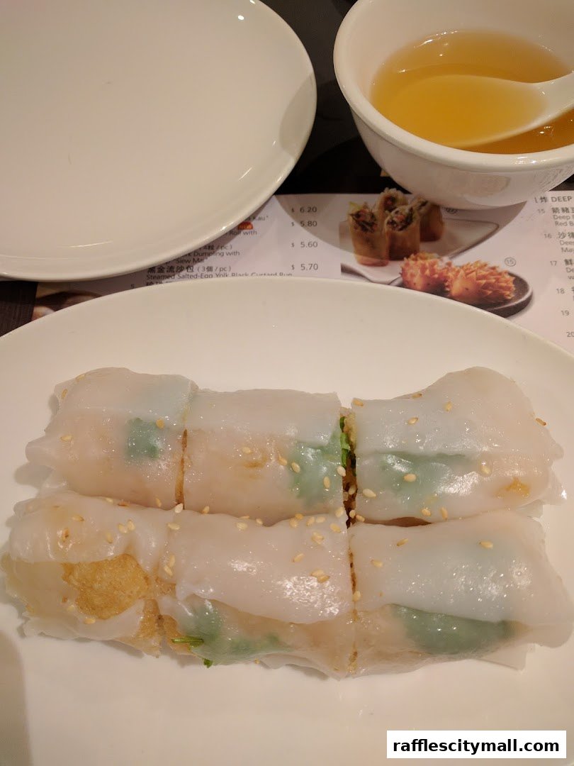 A plate of steamed rice noodle rolls with a small bowl of soup and a menu in the background.