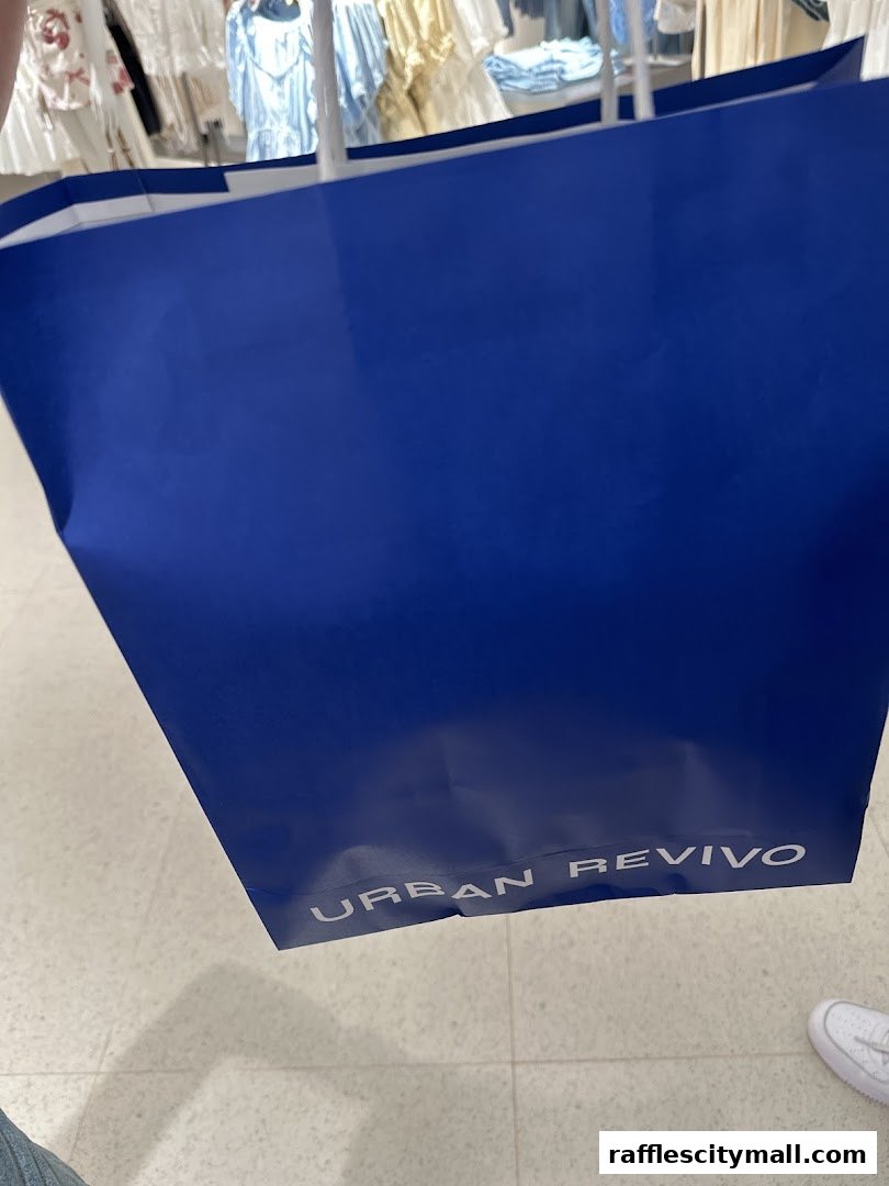 A blue shopping bag with the URBAN REVIVO logo, held in front of a clothing store display.