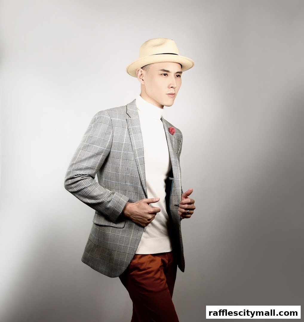 A man wearing a plaid blazer, turtleneck, and fedora poses against a grey background.