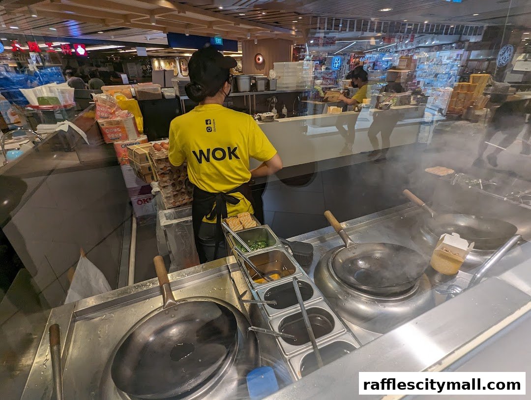 A chef in a yellow WOK HEY shirt prepares food in a busy kitchen with woks and ingredients.
