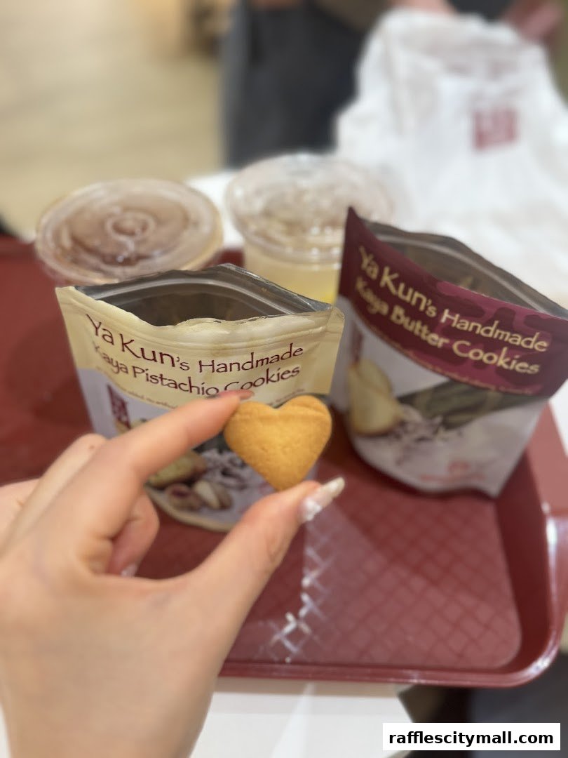 A hand holds a heart-shaped cookie with Ya Kun's cookies and drinks in the background.