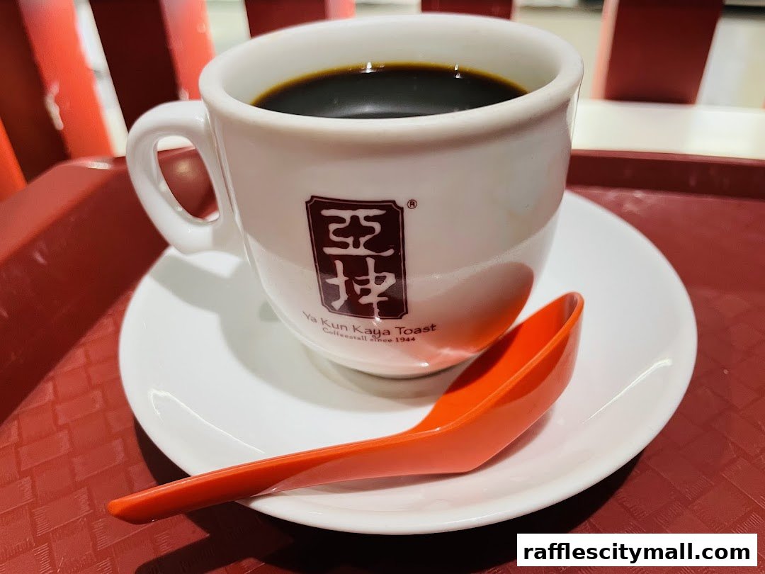 A close-up of a cup of black coffee with the Ya Kun Kaya Toast logo and an orange spoon.