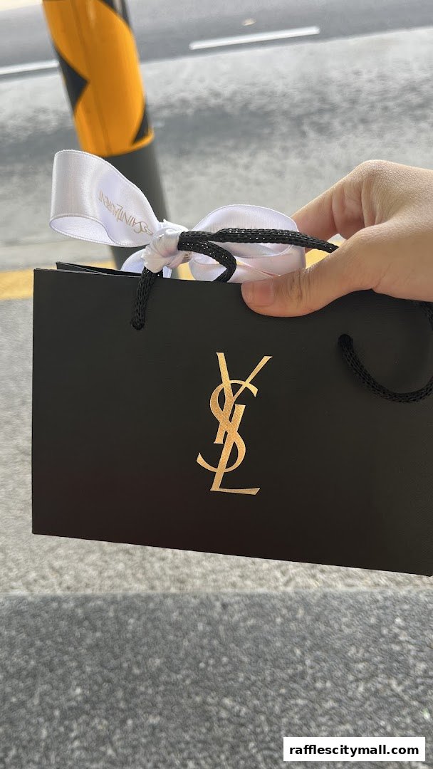 A hand holds a black YSL shopping bag with a gold logo and white ribbon.
