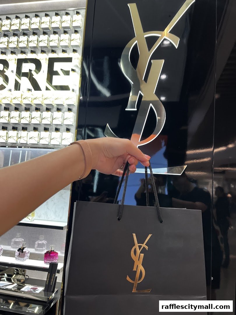 A hand holds a YSL shopping bag in front of a display of YSL products.